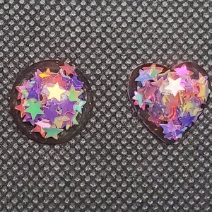May include: Five resin shapes filled with iridescent star-shaped glitter. The shapes are a circle, square, teardrop, heart, and a rounded rectangle.