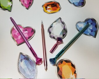 Puede incluir: Cuatro bolígrafos metálicos con tapas de joya en rosa, oro rosa, azul verdoso y azul. Los bolígrafos están sobre una superficie blanca con pegatinas de gemas de colores.