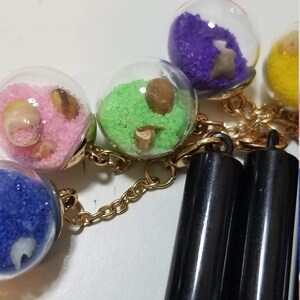 May include: A gold chain with five glass spheres containing different colored sand and small seashells. The chain is attached to two black pen-shaped objects.