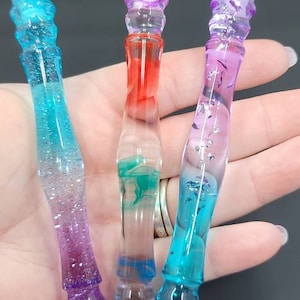 May include: Three glass pens with colorful swirls of blue, green, and purple. The pens are clear and have a decorative top.