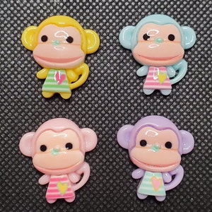 May include: Four plastic monkey figurines with different colored bodies and pink faces. Each monkey is wearing a striped shirt with a heart design. The monkeys are yellow, blue, pink, and purple.