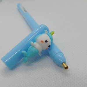 May include: A light blue pen with a blue and white narwhal charm attached to the top. The pen has a gold tip.