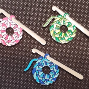 May include: Four enamel pins featuring a crochet hook and a crocheted circle. The circles are pink and white, green and yellow, blue and green, and blue and white.
