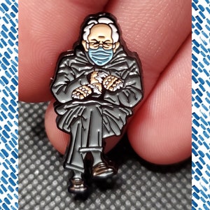 May include: Enamel pin featuring a seated figure in a winter coat, glasses, and a face mask. The figure is holding their arms and hands in front of them. The pin has a black outline with gray and tan coloring. The pin is approximately 1 inch in height.