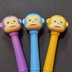 Może przedstawiać: Trzy długopisy w kształcie małpy o różnych kolorach korpusów: fioletowym, niebieskim i pomarańczowym. Każdy długopis ma małpią twarz z czarnymi oczami, czerwonym ustami i żółtym nosem.