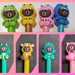 May include: Set of ten colorful pens with brown bear character toppers. The toppers are dressed in different animal costumes, including a frog, a giraffe, a pig, and a dinosaur.