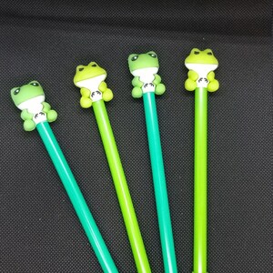 May include: Four pens with frog toppers in varying shades of green. The pens have a cylindrical design with a frog figure at the top. The frog figures are light green with white accents. The pens are arranged diagonally against a black background.
