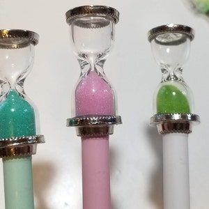 May include: Three miniature hourglass pens with different colored sand. The pens are teal, pink, and white.