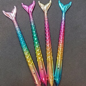 May include: Four mermaid tail pens with iridescent rainbow colors. The pens have a metallic finish and are arranged in a row.
