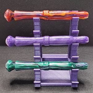 Könnte beinhalten: Drei Holzstifte mit verschiedenen marmorierten Mustern in verschiedenen Farben, die auf einem violetten Ständer präsentiert werden. Die Stifte sind rot, lila und grün.