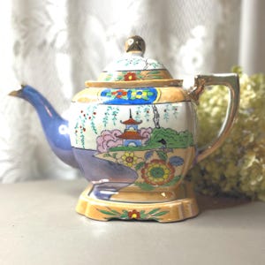 May include: A porcelain teapot featuring a hand-painted landscape with a pagoda, willow trees, and floral motifs in shades of blue, orange, yellow, and green. The teapot has a gold-trimmed handle, a matching lid with a gold finial, and a base.