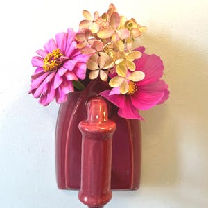 May include: A deep red ceramic wall-mounted vase shaped like a vintage iron, filled with a vibrant arrangement of pink zinnia flowers, a bright pink cosmos, and a cluster of dried, pale beige hydrangea blossoms.