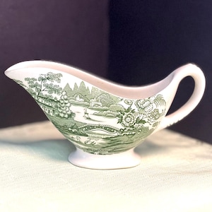 May include: A white ceramic gravy boat featuring a green transferware pattern depicting a landscape with a bridge, trees, buildings, and swans. The vessel has a curved handle, a wide pouring spout, and a pedestal base, showcasing a detailed scene.