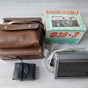 May include: A brown leather camera case, a small black leather accessory pouch, a vintage cardboard box labeled "ФЭД-3" with a camera illustration, and a grey rectangular electronic device with a white power cord and a black front panel.