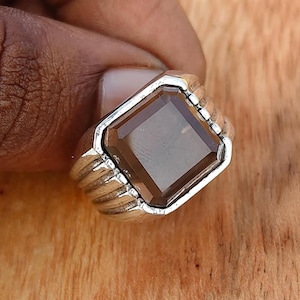 May include: A silver-toned ring featuring a large, square-cut, translucent brown gemstone set in a polished metal band. The band has a ribbed, multi-grooved design on the sides, leading up to the central stone setting, creating a structured appearance.
