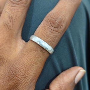 Op de afbeelding: Een eenvoudige, gladde, gepolijste zilverkleurige bandring met een plat, uniform oppervlak. De ring heeft een strak ontwerp met een reflecterende metalen afwerking die het licht opvangt en de vinger omsluit met een consistente breedte.