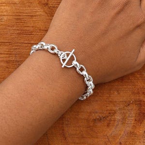 May include: A silver-toned chain link bracelet featuring a toggle clasp closure. The bracelet is constructed from a series of polished, rounded metal links that connect to a central circular ring and a perpendicular bar mechanism for fastening.