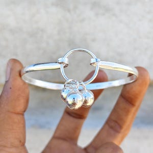May include: A silver-toned bangle bracelet featuring a central circular ring component. Three small, smooth, spherical silver-toned charms are attached to the bottom of the central ring, hanging freely from the main structure of the bracelet.
