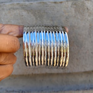 Puede incluir: Un conjunto de diez brazaletes de metal en tono plateado pulido con un perfil liso y redondeado. Las pulseras están apiladas, creando una apariencia uniforme y reflectante con un acabado de alto brillo que capta la luz.