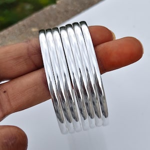 以下が含まれることがあります： 滑らかで丸みを帯びた表面を持つ、6本の磨かれたシルバー調のバングルセット。各バングルは光を反射する金属仕上げが施されており、円形のブレスレットを重ねた状態で均一な外観を作り出しています。
