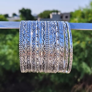 May include: A set of ten silver-toned metal bangles arranged in a row. Each bangle features a distinct embossed pattern, including floral motifs, braided textures, wavy lines, and geometric shapes, all with a polished, reflective metallic finish.
