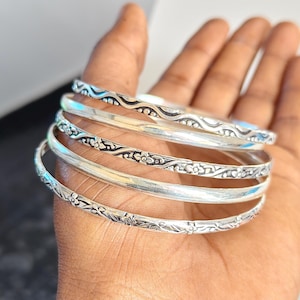 Può includere: Un set di cinque braccialetti rigidi color argento caratterizzati da un mix di superfici lisce e lucide e intricati motivi floreali o ondulati in rilievo. I bracciali sono impilati, mostrando texture e design vari con finitura argento.
