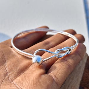 Puede incluir: Una pulsera rígida de plata pulida con una banda metálica lisa y redondeada que se curva en un diseño de nudo decorativo. Un extremo de la pulsera termina en una pequeña esfera de plata, mientras que el otro extremo forma un cierre de bucle.