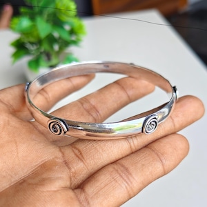 Può includere: Un bracciale rigido color argento lucido caratterizzato da una fascia liscia e arrotondata, decorata con motivi a spirale circolari distanziati uniformemente. La superficie metallica ha una finitura riflettente e i dettagli a spirale sono messi in risalto da linee scure.