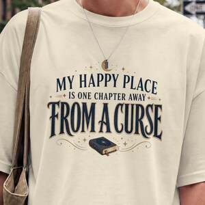 Op de afbeelding: Een off-white t-shirt met korte mouwen en donkerblauwe tekst met de tekst "MY HAPPY PLACE IS ONE CHAPTER AWAY FROM A CURSE", met een kleine illustratie van een gesloten boek en decoratieve sterren in het midden op de borst.