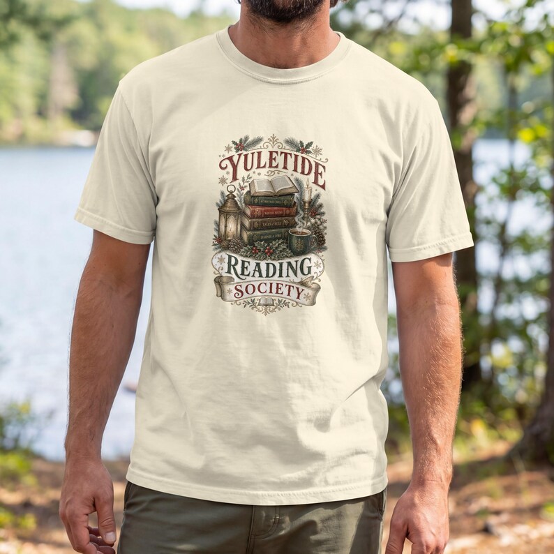 May include: A cream-colored t-shirt featuring a graphic print of a stack of books, a lantern, a candle, and a mug. The text reads "YULETIDE READING SOCIETY" in a decorative font, surrounded by pine branches, holly berries, and snowflakes.