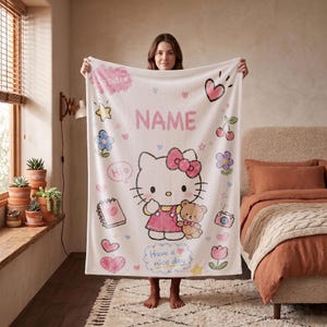 May include: A white fleece blanket featuring a central graphic of Hello Kitty holding a teddy bear, surrounded by colorful illustrations of hearts, flowers, a notebook, a camera, and cherries, with the text NAME and Have a nice day printed.