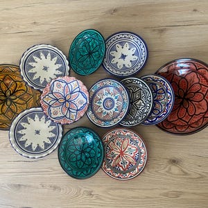 May include: A collection of twelve round ceramic plates and bowls featuring intricate hand-painted geometric and floral patterns in vibrant colours including turquoise, mustard yellow, terracotta, navy blue, and white with scalloped edges.