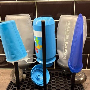 May include: A black drying rack with a hexagonal grid base and vertical posts holding three hydration flasks and a blue cap. The flasks are translucent and blue, with one featuring the text ULTIMATE DIRECTION printed in white on the side.