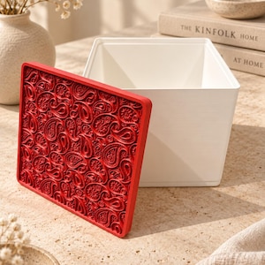 Può includere: Un contenitore quadrato bianco con apertura superiore e un coperchio rosso separato. Il coperchio presenta un motivo paisley complesso e in rilievo su tutta la superficie. Il contenitore è realizzato in materiale liscio e solido con bordi.