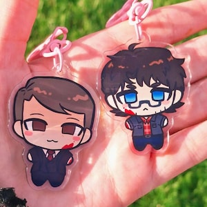 May include: Two clear acrylic keychains featuring chibi-style character illustrations. One character wears a dark suit with a red tie, and the other wears a dark jacket over a red plaid shirt with blue glasses. Both figures have red blood-like marks.