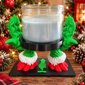 May include: A clear glass candle jar with light blue wax sits on a black circular holder supported by two green legs with white fur-trimmed cuffs and red feet. A small green silhouette of a character is centred on the black base.