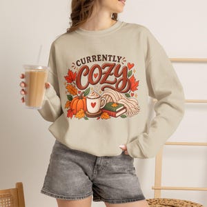 May include: A beige crewneck sweatshirt featuring a graphic print with the text "CURRENTLY COZY" in brown, accompanied by illustrations of a pumpkin, a mug with a heart, autumn leaves, a stack of two books, and a knitted scarf in warm tones.