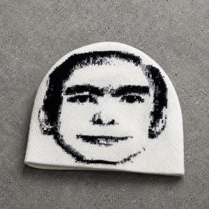 May include: A white knit beanie featuring a high-contrast black graphic print of a human face. The design displays a stylized, pixelated portrait of a person with dark hair, prominent eyebrows, and a neutral expression centered on the front of the cap.
