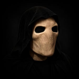 Op de afbeelding: Een masker voor het hele hoofd met een glad, beige, gelaatloos gezicht met diepe, donkere, holle oogkassen. Het masker is geïntegreerd met een zwarte stoffen capuchon die over het hoofd en de schouders valt en een scherp contrast vormt.