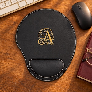 May include: A black leather mouse pad with a built-in wrist rest, featuring white contrast stitching along the edges and a gold-colored ornate letter A monogram printed in the center of the surface for a decorative and functional desk accessory.