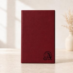 May include: A rectangular, deep burgundy notebook with a smooth, solid-colored cover. The bottom right corner features a black, ornate, calligraphic letter A surrounded by delicate, swirling flourishes and decorative scrollwork patterns.