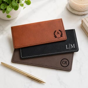 May include: Three rectangular checkbook covers in tan, black, and dark gray. The tan cover features a laurel wreath with the letter A. The black cover has white stitching and the letters L|M. The gray cover displays the letters CR in a circle.