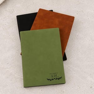 May include: Three rectangular notebooks stacked together. The top notebook is olive green with the text "SALLY GARRISON SH" and a small floral graphic. Beneath it are a brown notebook and a black notebook, all featuring a smooth, matte finish.