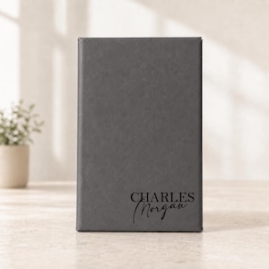 May include: A rectangular dark gray notebook with a smooth, leather-like cover. The name "CHARLES Morgan" is printed in black on the bottom right corner, featuring a combination of bold sans-serif and elegant script typography.