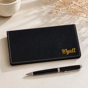 May include: A black textured checkbook cover with white contrast stitching and the name Wyatt embossed in gold script on the front, accompanied by a sleek black ballpoint pen with silver accents and a silver clip positioned below the cover.