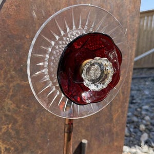 May include: A decorative garden stake featuring a layered flower design made from stacked glass elements. The base is a clear, starburst-patterned glass plate, topped with a smaller, deep red glass bowl, and centered with a clear, faceted glass knob.