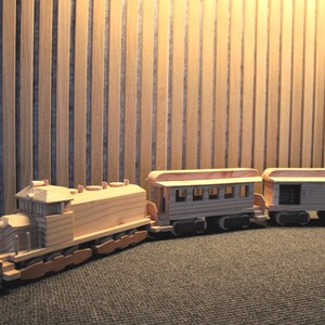Könnte beinhalten: Ein hölzernes Spielzeugeisenbahn-Set, bestehend aus einer Lokomotive und zwei Personenwagen. Der Zug ist aus hellem Naturholz mit sichtbarer Maserung gefertigt und verfügt über kleine Räder sowie detaillierte, rechteckige Fensterausschnitte.