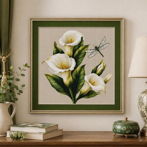 May include: A square framed wall art piece featuring a botanical illustration of three white calla lilies with green leaves and a dragonfly. The artwork is set against a light beige background and enclosed in a dark green mat and a gold-toned frame.
