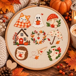 May include: A circular wooden embroidery hoop containing a cross-stitch sampler on cream fabric. The design features nine autumn-themed motifs: a cat in a plaid blanket, a ghost holding a pumpkin, a mushroom, a house, a heart-shaped wreath, an umbrella, a hedgehog, a paper airplane, and a pair of socks.