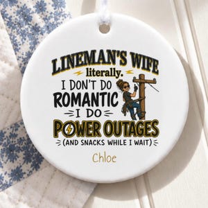 May include: A round white ceramic ornament featuring a cartoon illustration of a lineman on a utility pole and the text: LINEMAN'S WIFE literally. I DON'T DO ROMANTIC I DO POWER OUTAGES (AND SNACKS WHILE I WAIT) with the name Chloe below.